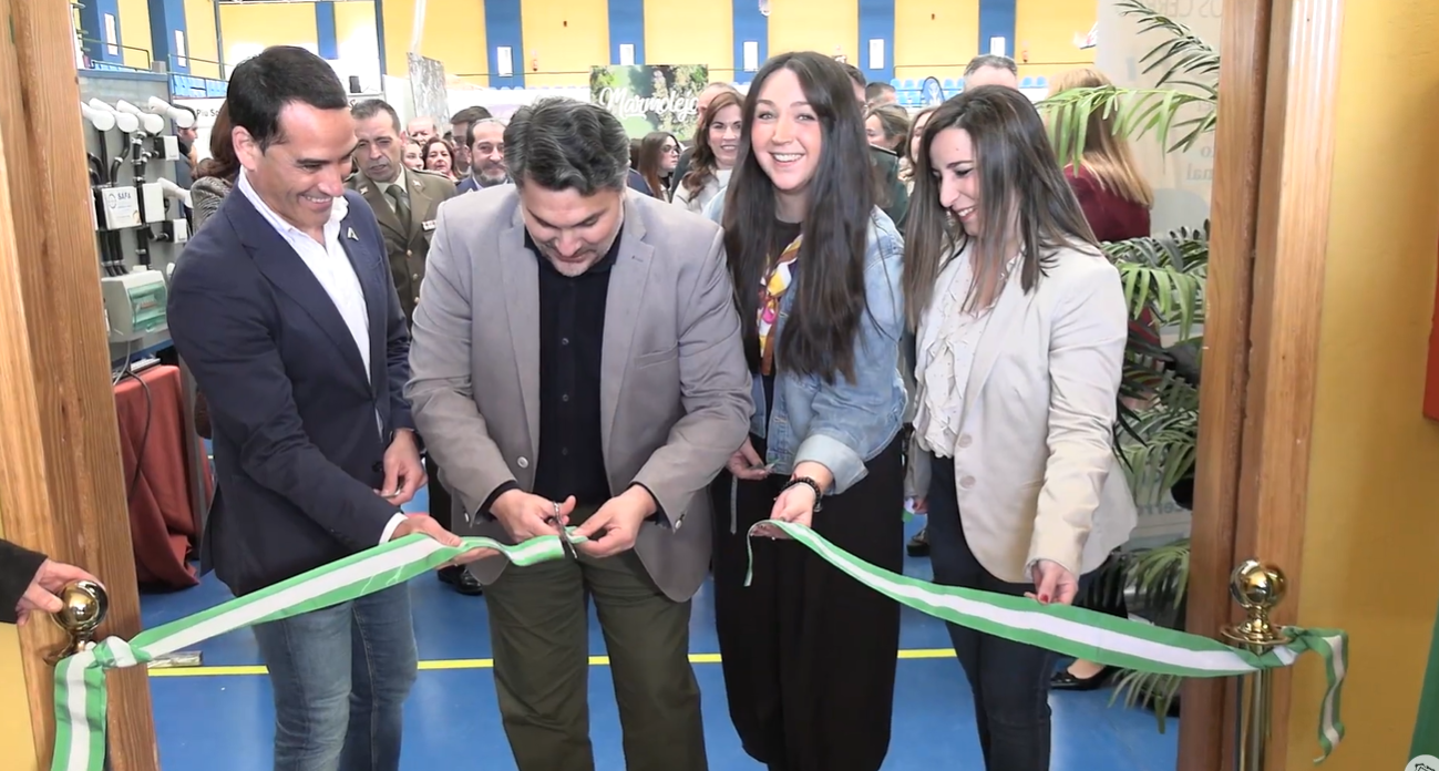 Inauguración del Salón X de la Juventud. Cuatro personas cortando una cinta verde en una sala con plantas, paredes azules y amarillas. La diputada María Dolores Ruiz en el centro junto a otros miembros