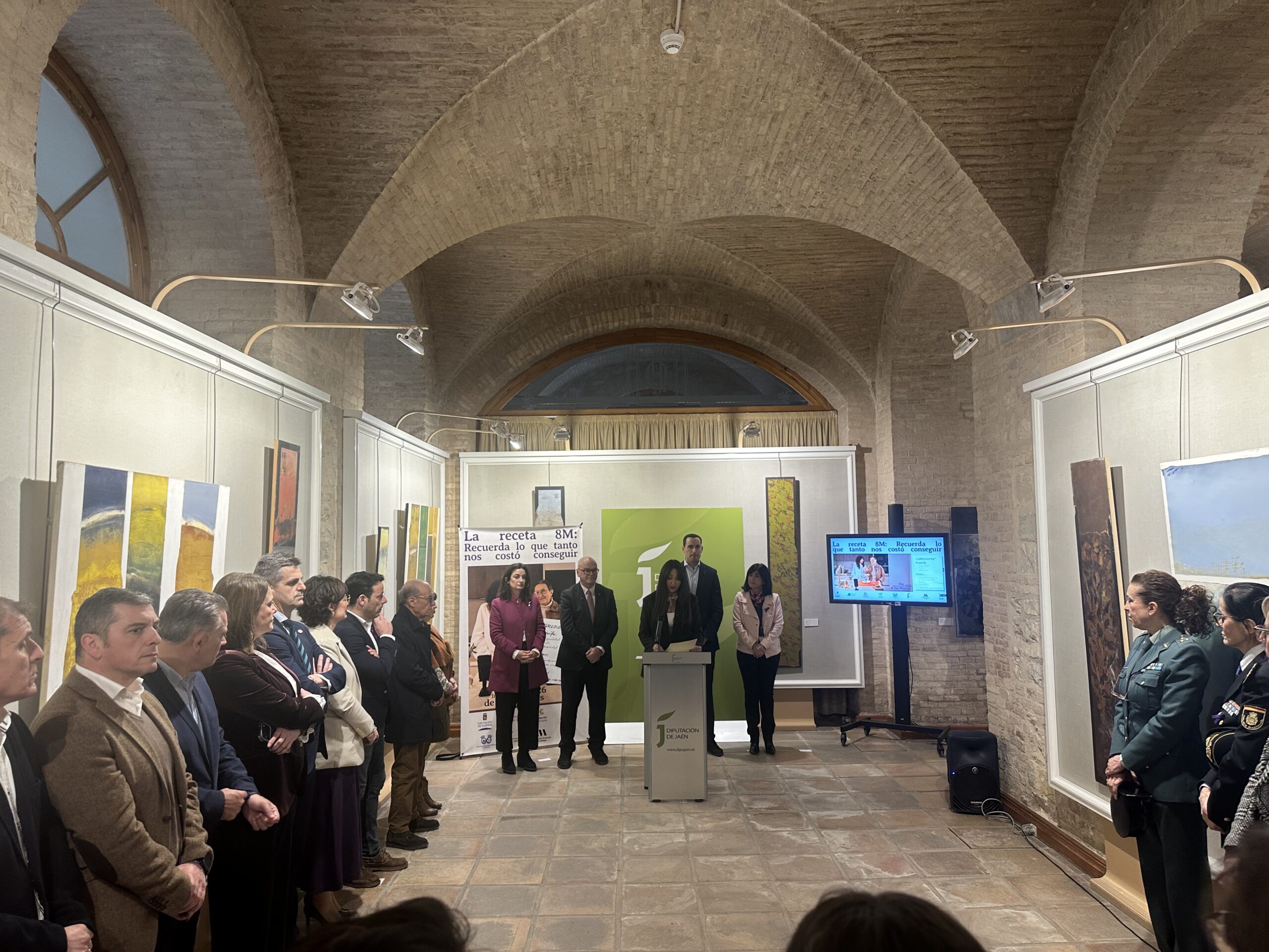 Presentación de las campañas de igualdad del 8M de la Diputación de Jaén en un aula con bóveda de ladrillo. Una persona habla desde un podio con el logo de la Diputación frente a un grupo de asistentes que observan atentamente.