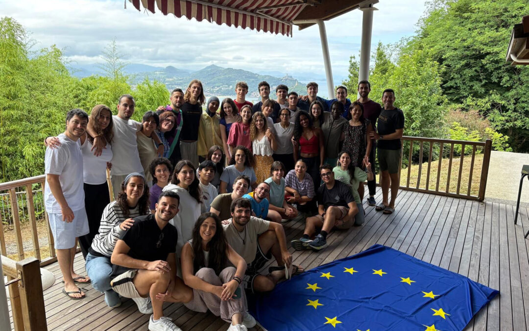 Grupo de jóvenes participantes en el Encuentro Juvenil de Técnicos Políticos sobre salud mental posando en una terraza en San Sebastián con la bandera de la Unión Europea.