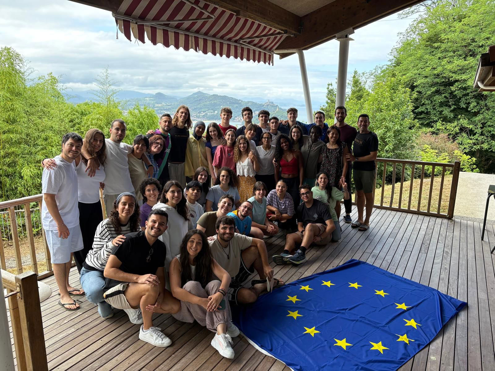 Grupo de jóvenes participantes en el Encuentro Juvenil de Técnicos Políticos sobre salud mental posando en una terraza en San Sebastián con la bandera de la Unión Europea.